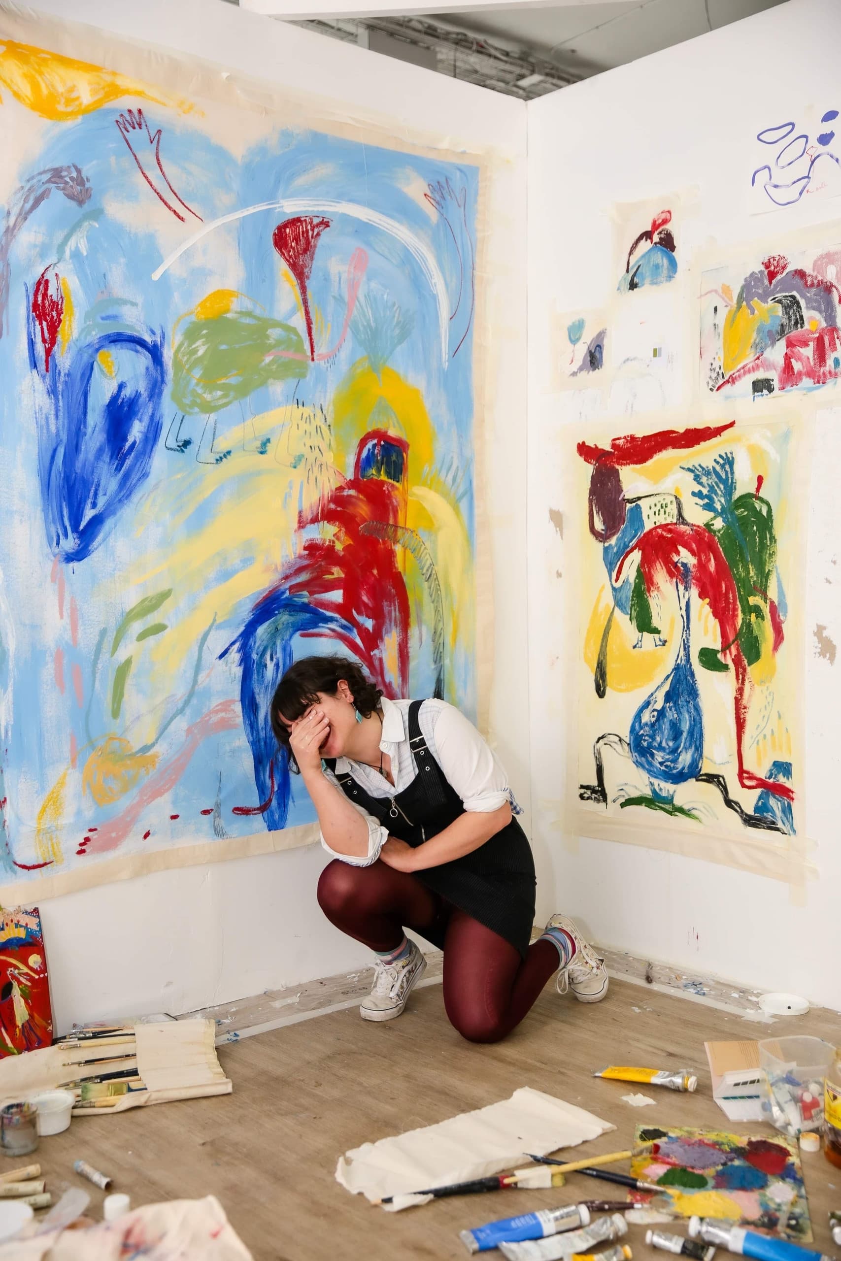Inês Miguel Oliveira kneeling in her studio, surrounded by colourful paintings on paper and painting materials.